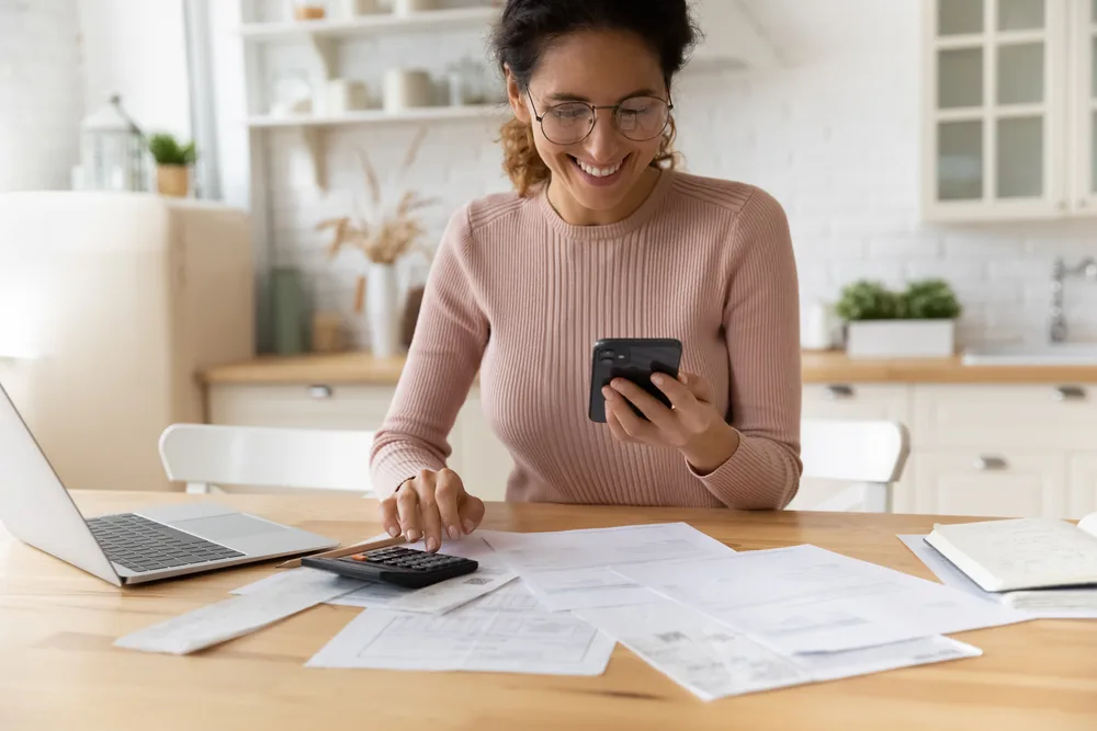 Woman reviewing bills with a calculator and phone at a kitchen table