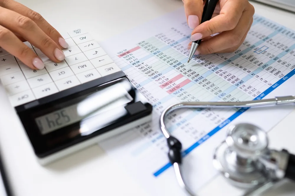 Hands using a calculator over a spreadsheet next to a stethoscope