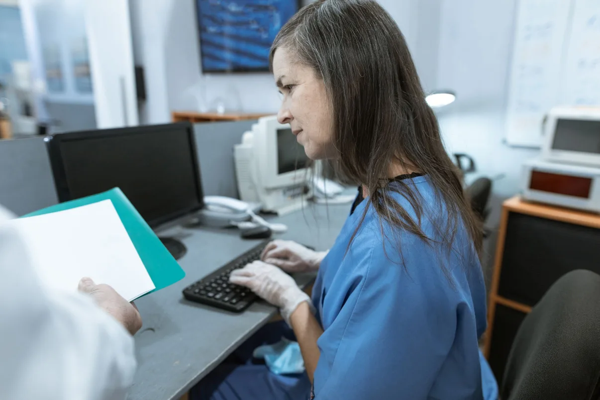 Medical billing specialist working on claims at a computer while a colleague brings paperwork